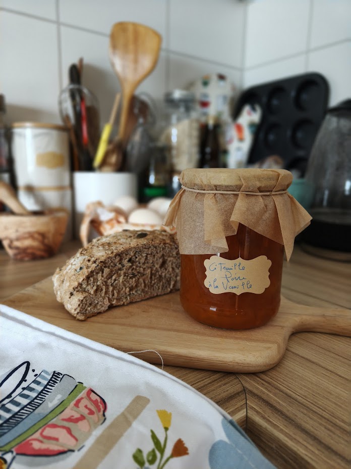 Jar of pumpkin, pear and vanilla jam under a paper cover, beside a slice of seeded bread