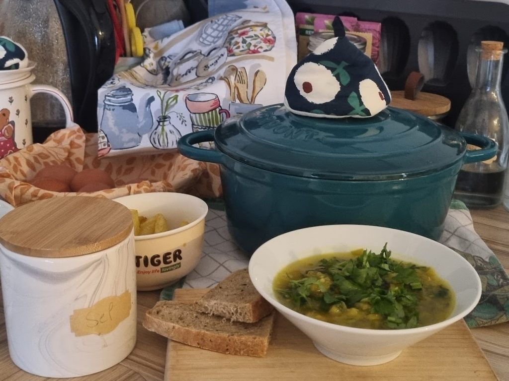 Bowl of soup with coriander in front of a teal casserole pot, bread on the board