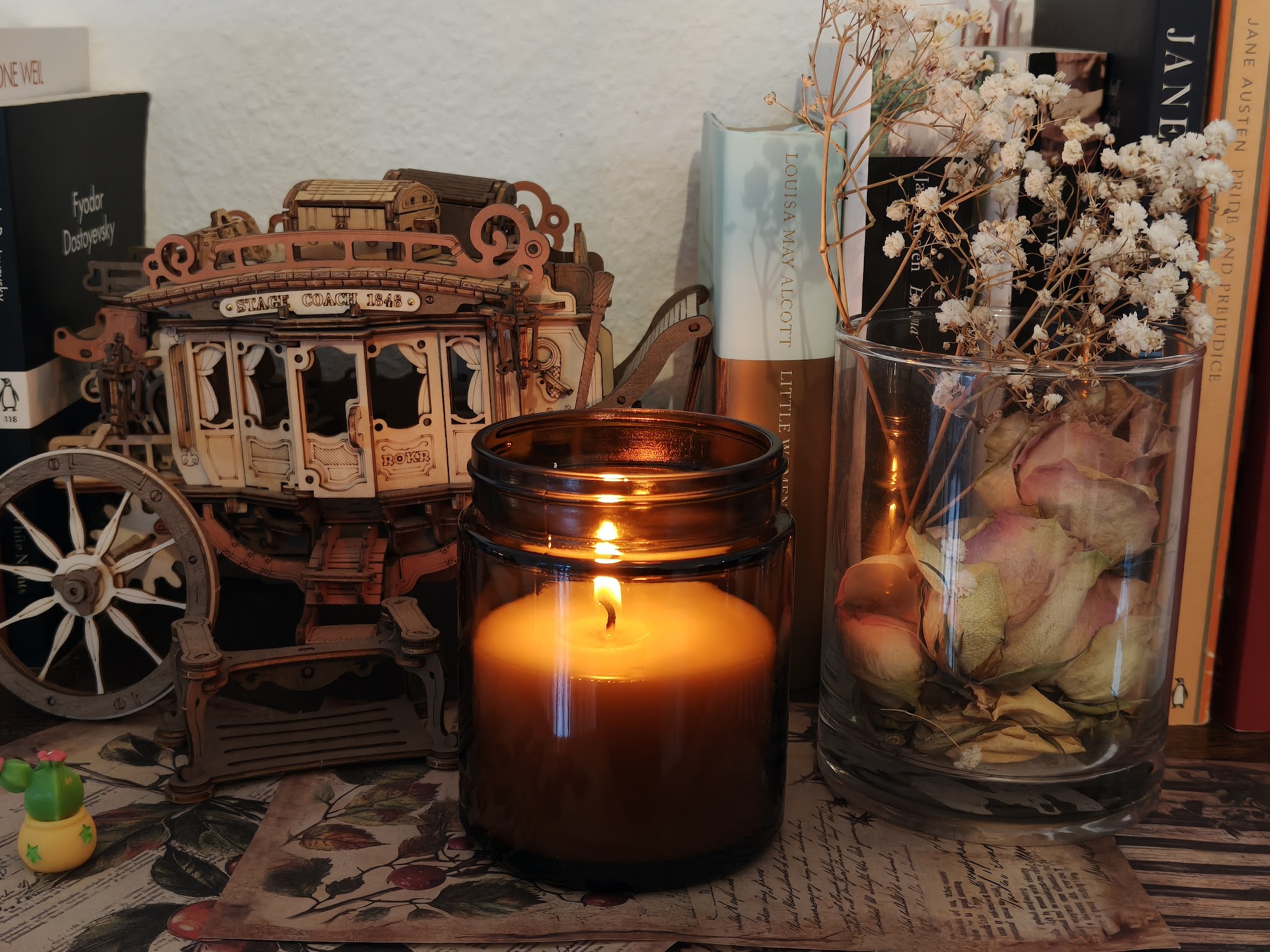 A lit candle in an amber jar beside a wooden stagecoach model, a jar of dried roses and a row of books