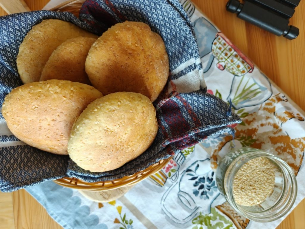 Basket of sesame-topped semolina rolls in a blue cloth, beside a jar of sesame seeds